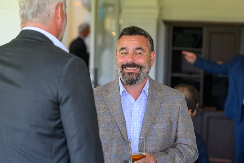 Homme souriant avec une barbe, tenant un verre, en costume à un événement.